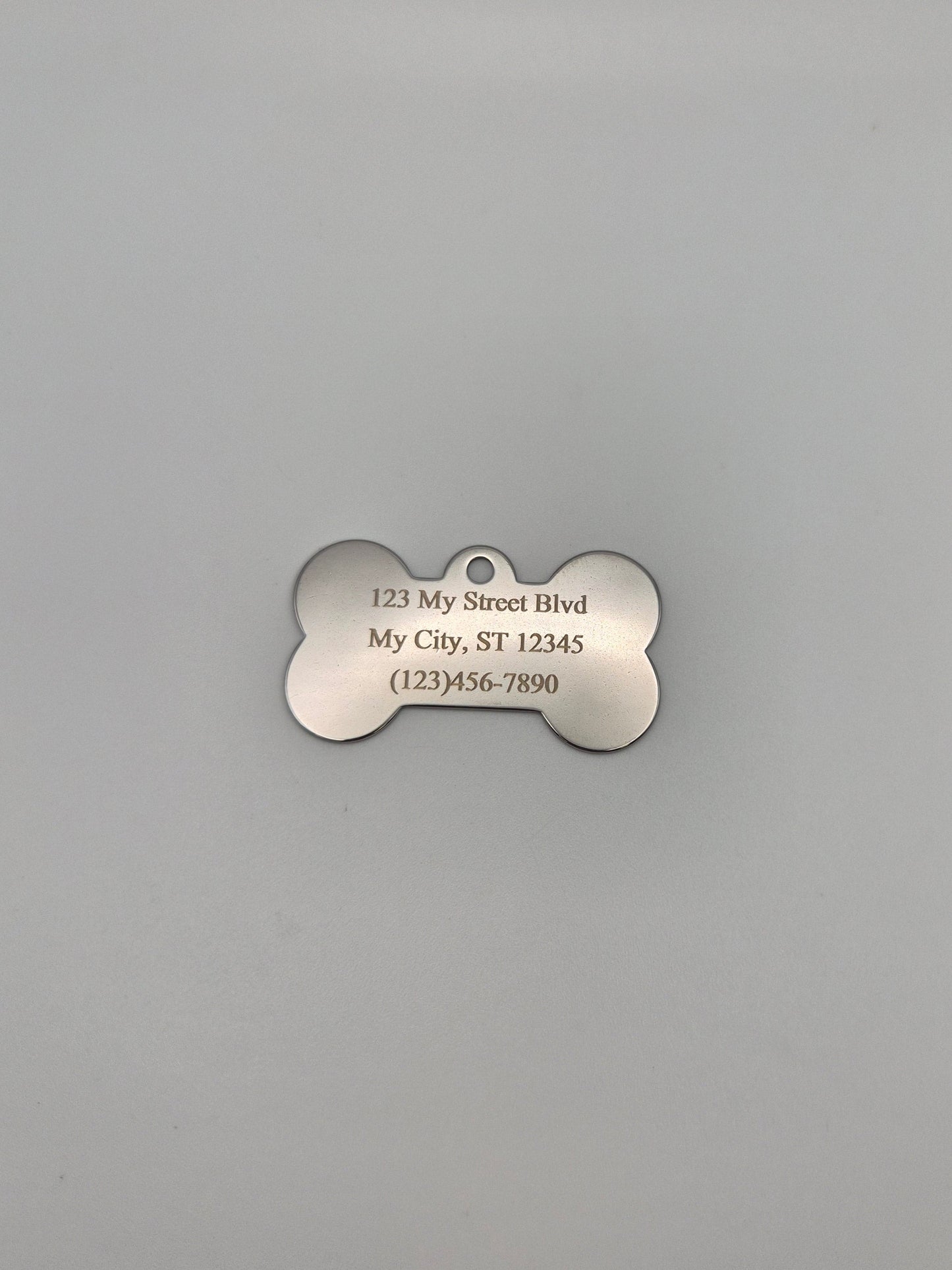 Stainless steel, bone-shaped dog tag with engraved full address and phone number on a gray background