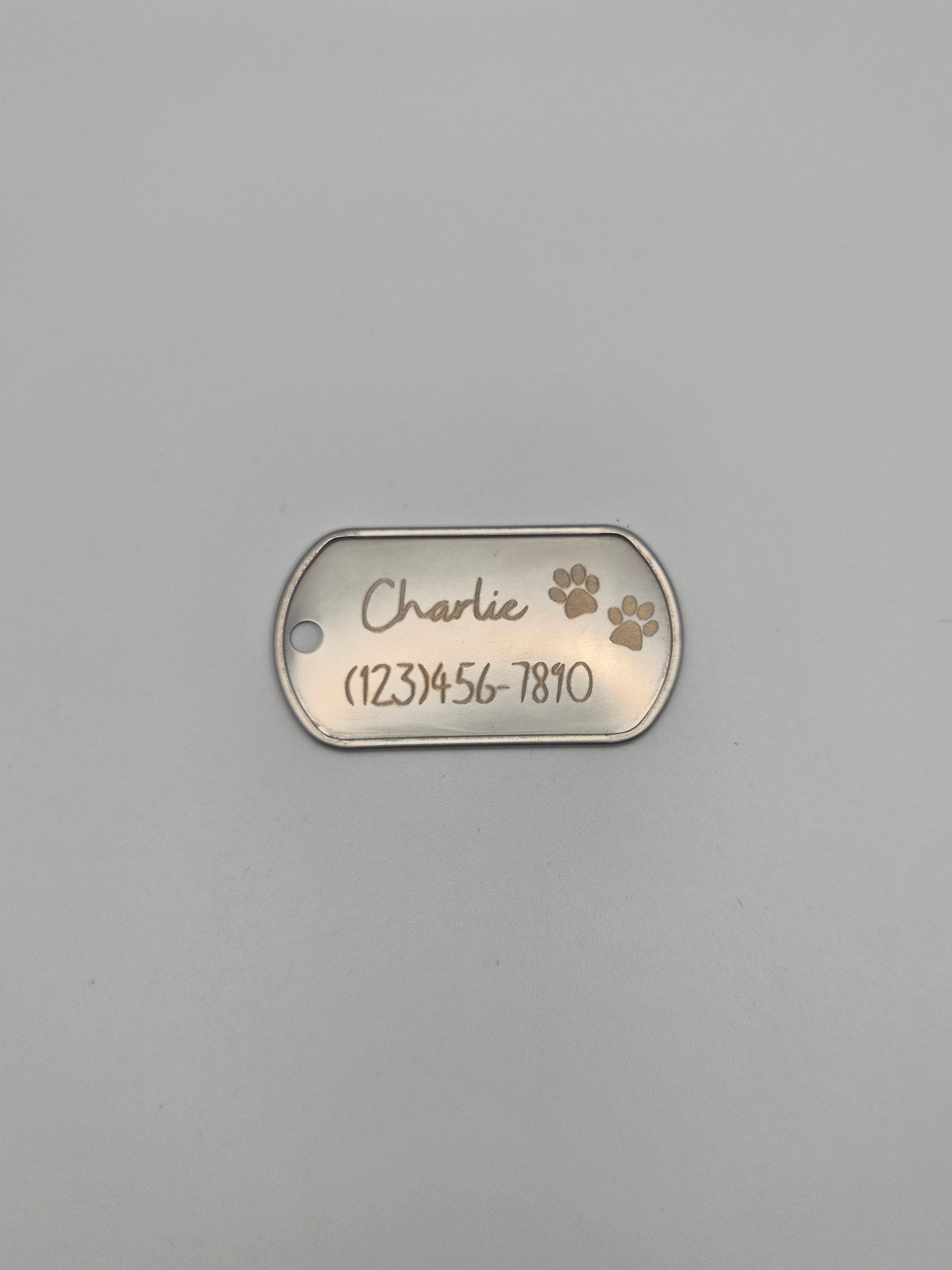 Metal dog tag with 2 engraved pawprints, the name 'Charlie,' and phone number on a gray background