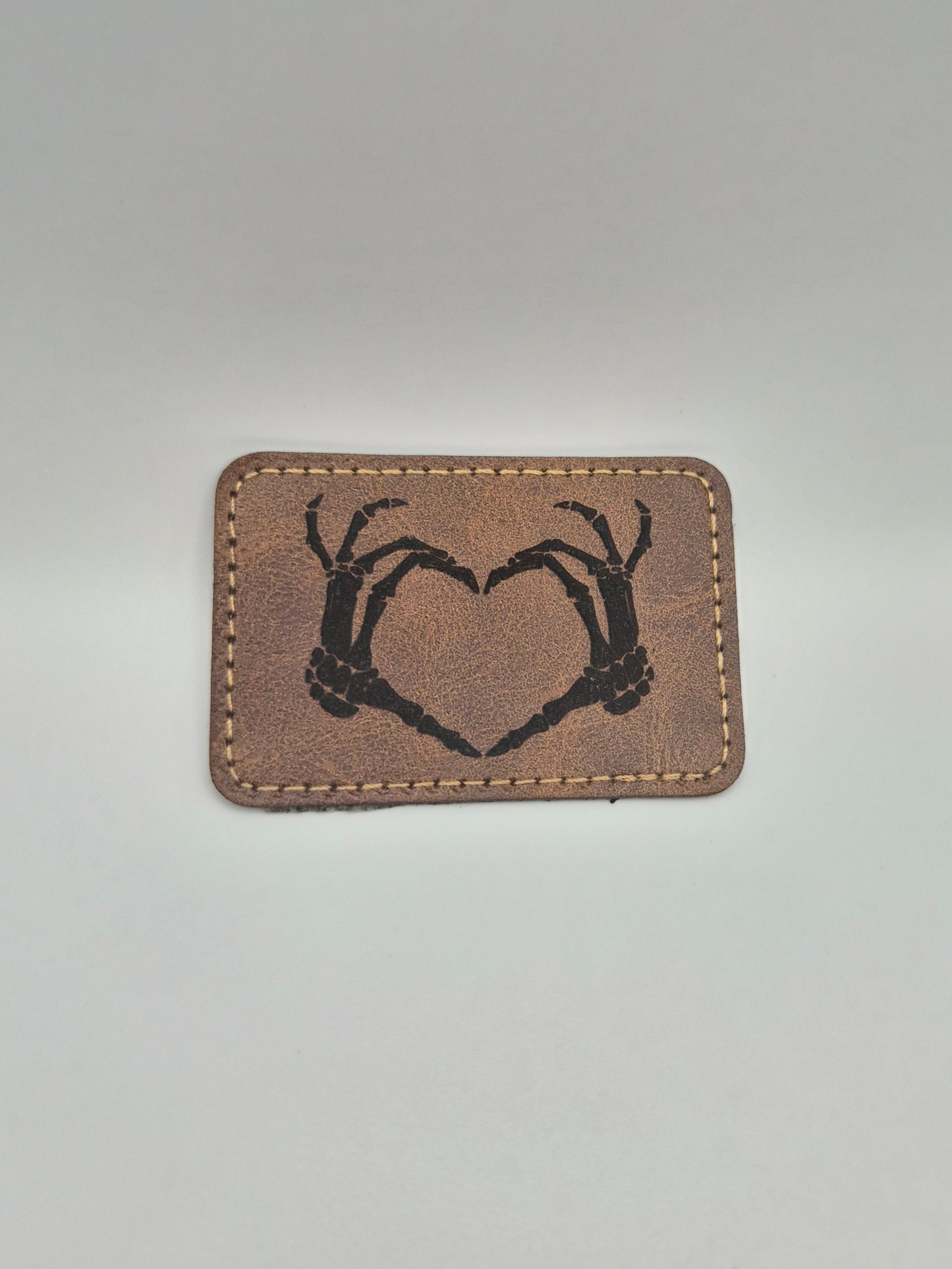 Brown leather patch with an engraved image of a heart created by 2 skeleton hands on a gray background
