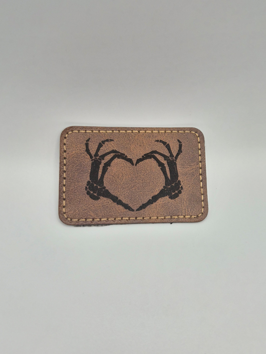 Brown leather patch with an engraved image of a heart created by 2 skeleton hands on a gray background
