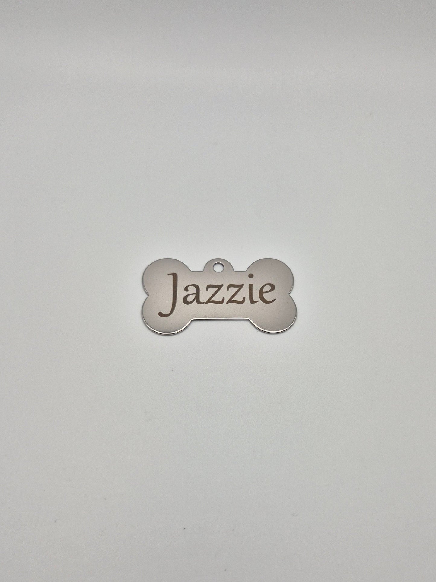 Stainless steel, bone-shaped dog tag with 'Jazzie' engraved on it, sitting on a light gray background