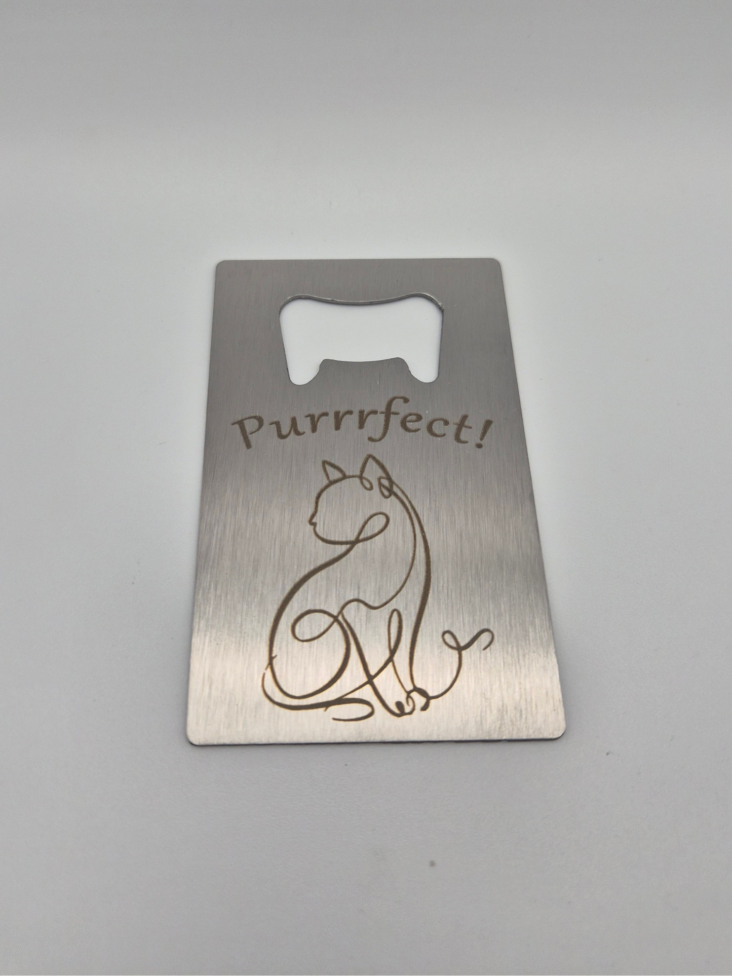 Brushed, stainless steel, credit card-sized bottle opener with an engraved single-line-drawing-style cat design and the text 'Purrfect!' sitting on a light background