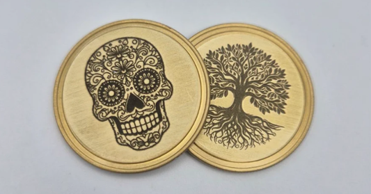Two round brass coins with engraved designs, one featuring a decorative sugar skull and the other a tree of life, displayed on a light gray background.