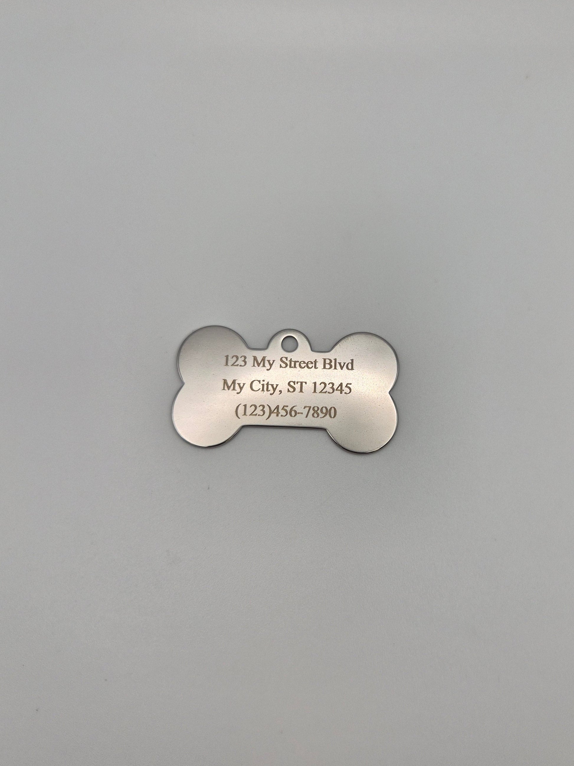 Stainless steel, bone-shaped dog tag with engraved full address and phone number on a gray background