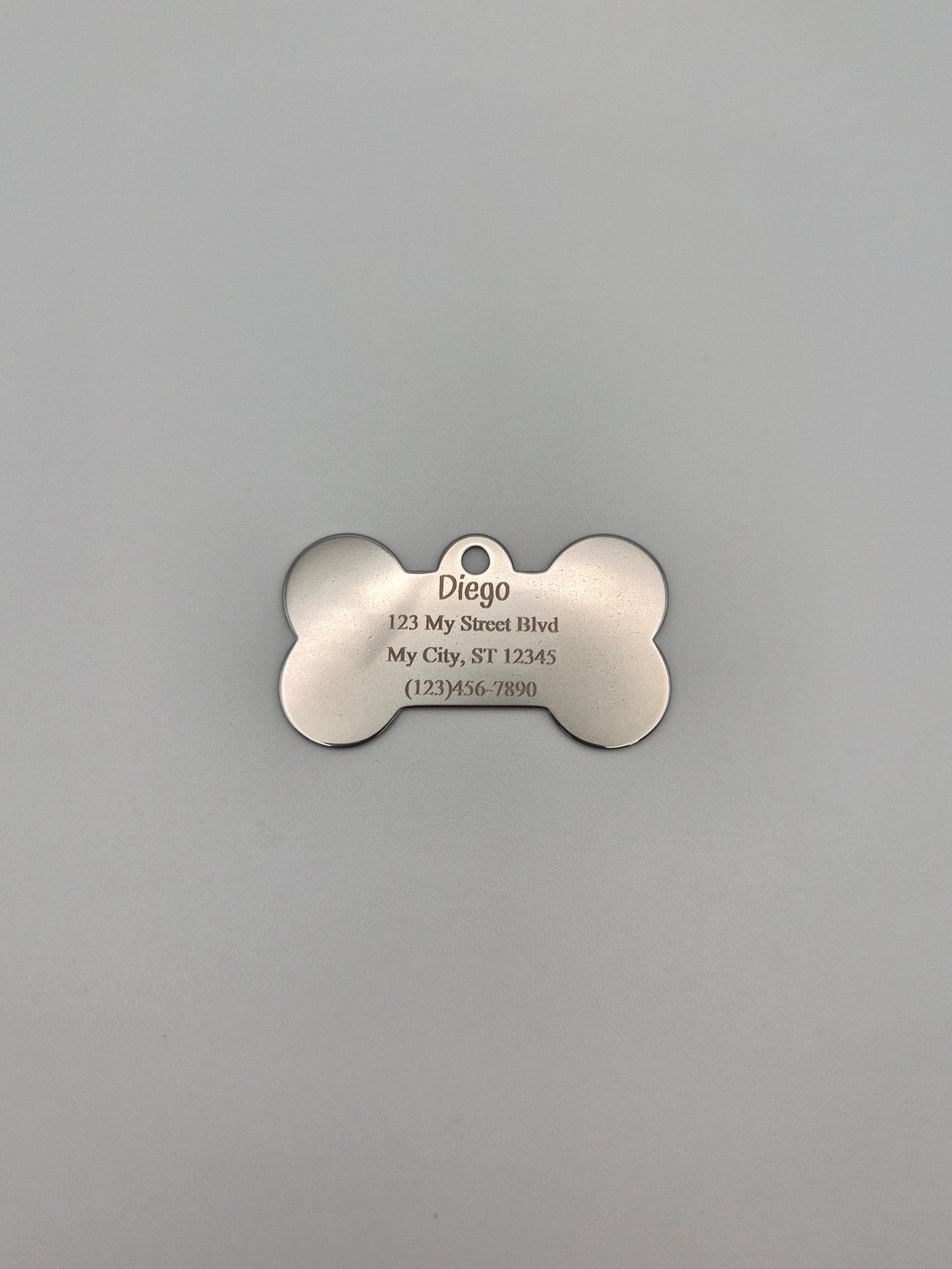 Stainless steel, bone-shaped dog tag with 4 lines of engraved text on a gray background