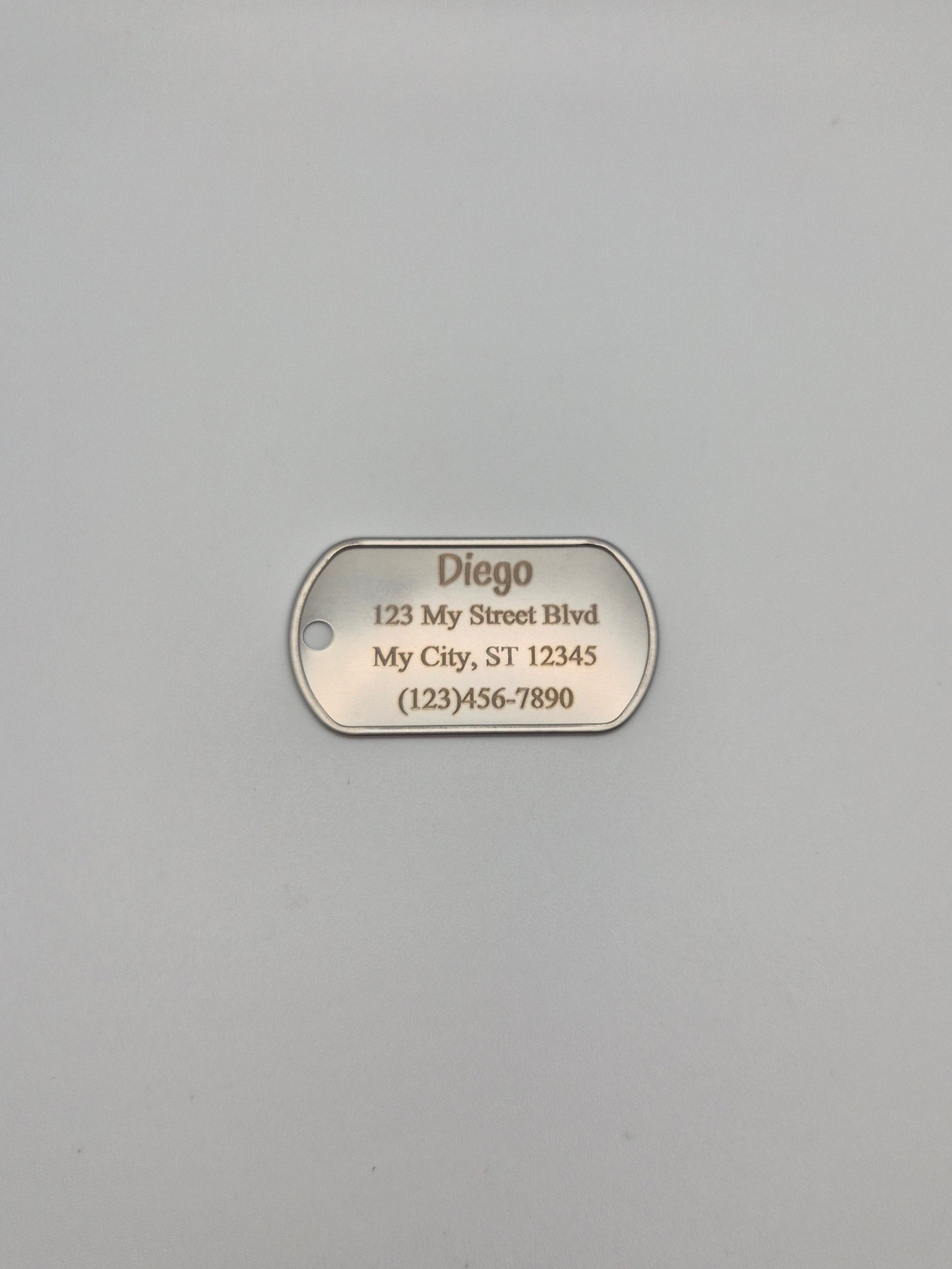 Aluminum military ID-style dog tag with engraved name, full address, and phone number sitting on a gray background
