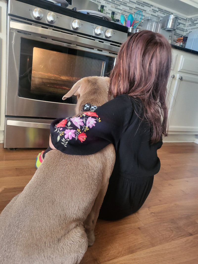 Person wearing a black hoodie with floral designs, standing next to a dog in a kitchen.