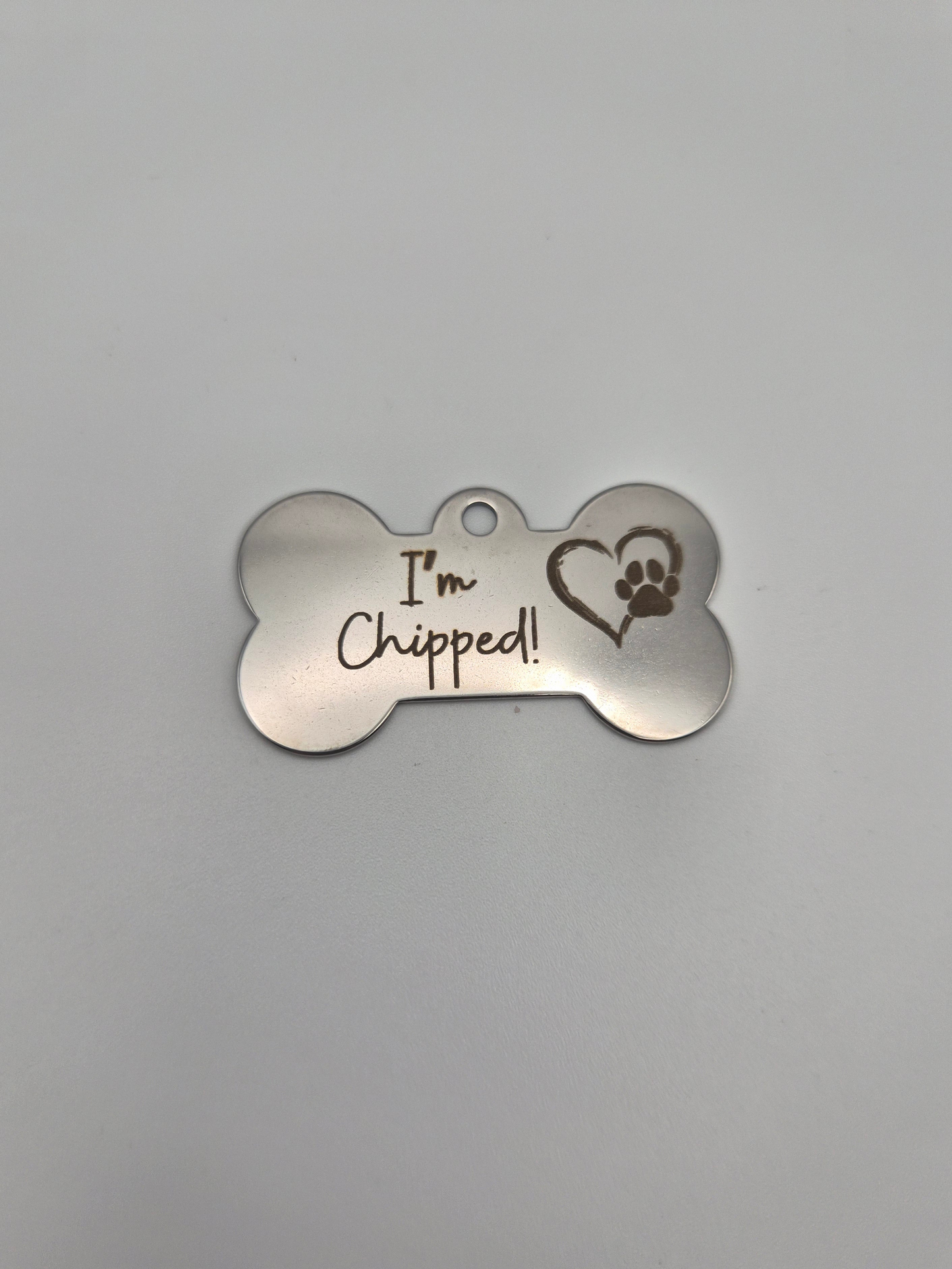 Stainless steel bone-shaped dog tag with 'I'm Chipped!' text and a heart-paw engraved on it sitting on a light background