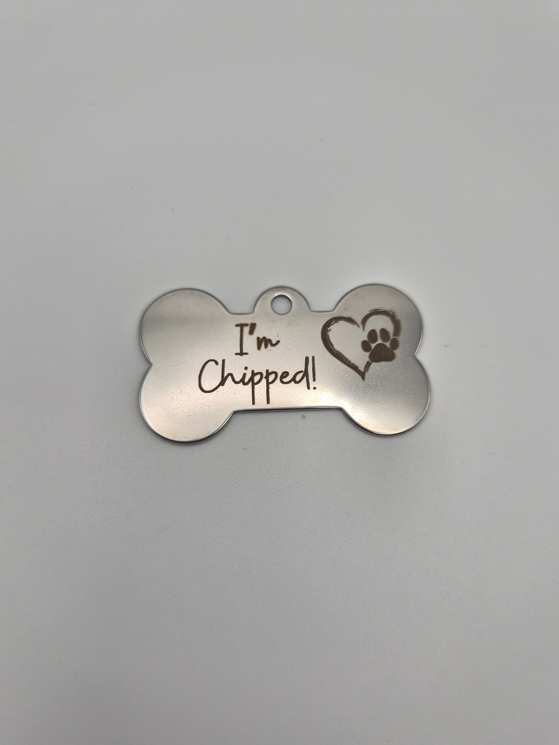 Stainless steel bone-shaped dog tag with 'I'm Chipped!' text and a heart-paw engraved on it sitting on a light background