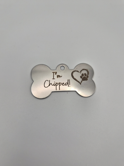 Stainless steel bone-shaped dog tag with 'I'm Chipped!' text and a heart-paw engraved on it sitting on a light background