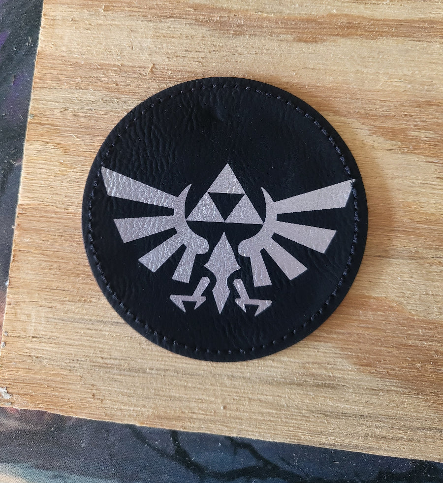 A circular black leather patch featuring the silver-gray Royal Crest from The Legend of Zelda, displayed on a light wood surface.