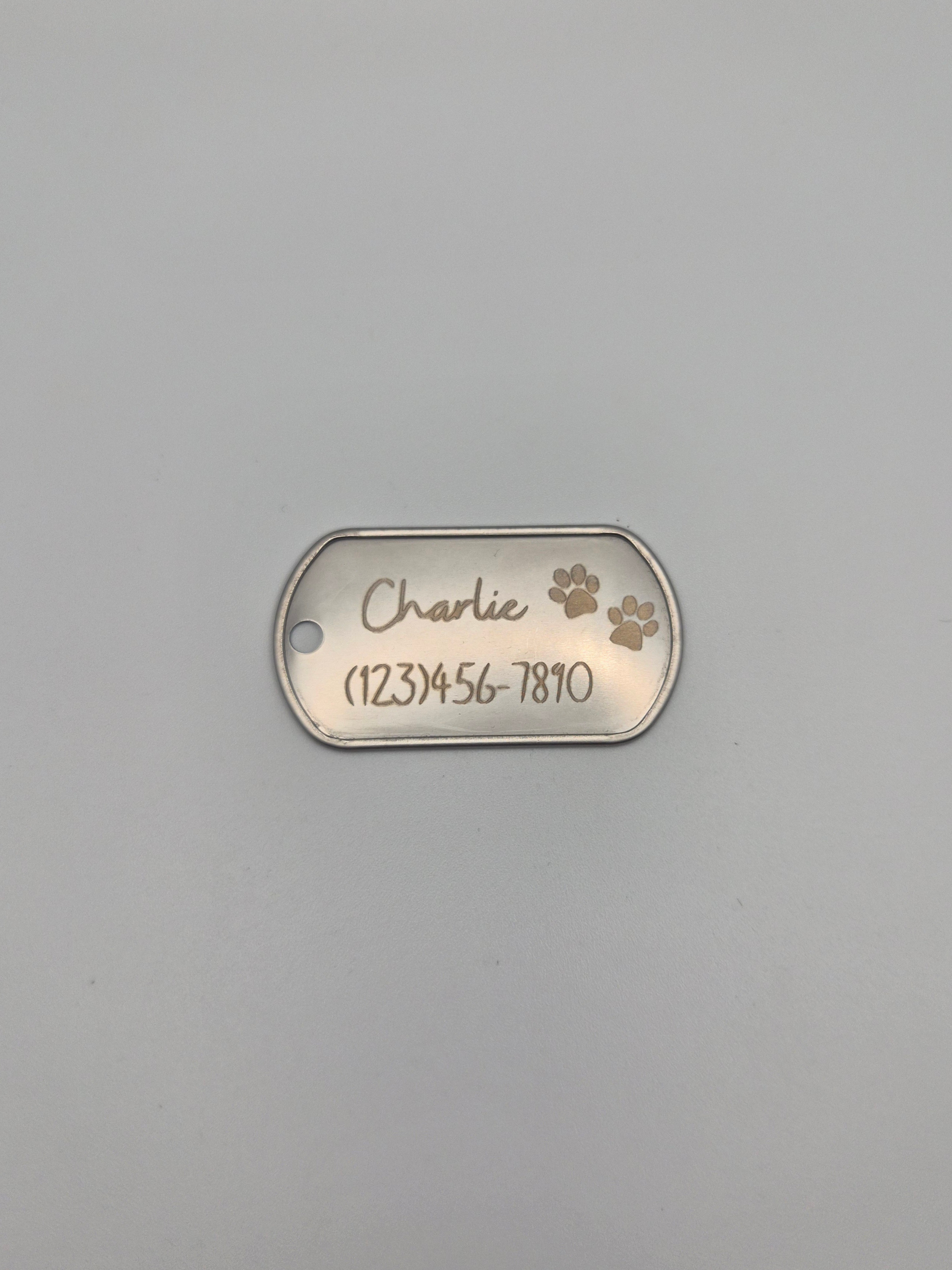 Metal dog tag with 2 engraved pawprints, the name 'Charlie,' and phone number on a gray background