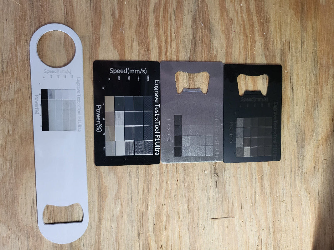 Two credt card bottle openers, one aluminum business card, and a white bar key-style bottle opener laid out on a wooden surface. Each item has been engraved with a test grid to show engraved color and speed/power results.