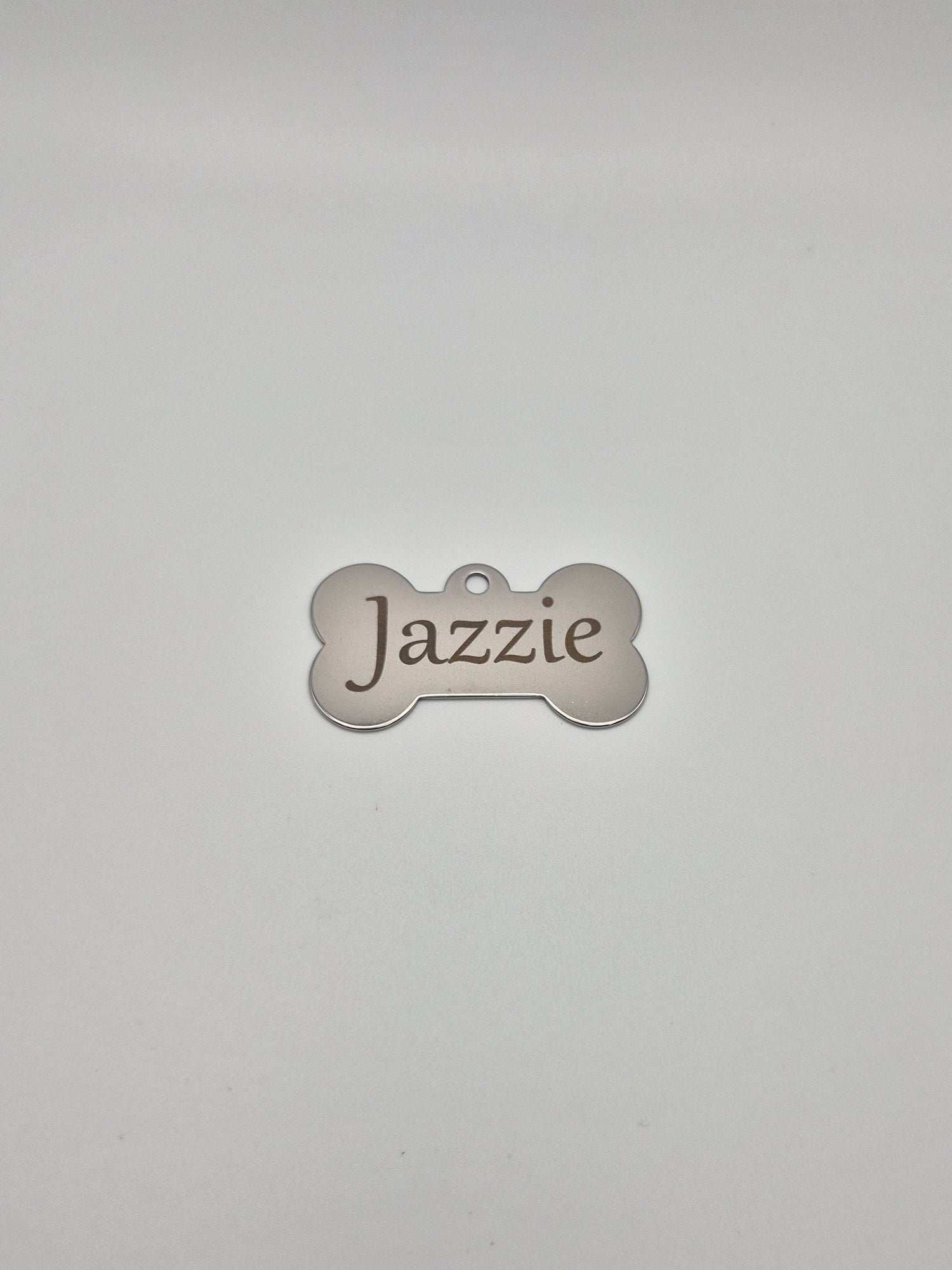 Stainless steel, bone-shaped dog tag with 'Jazzie' engraved on it, sitting on a light gray background