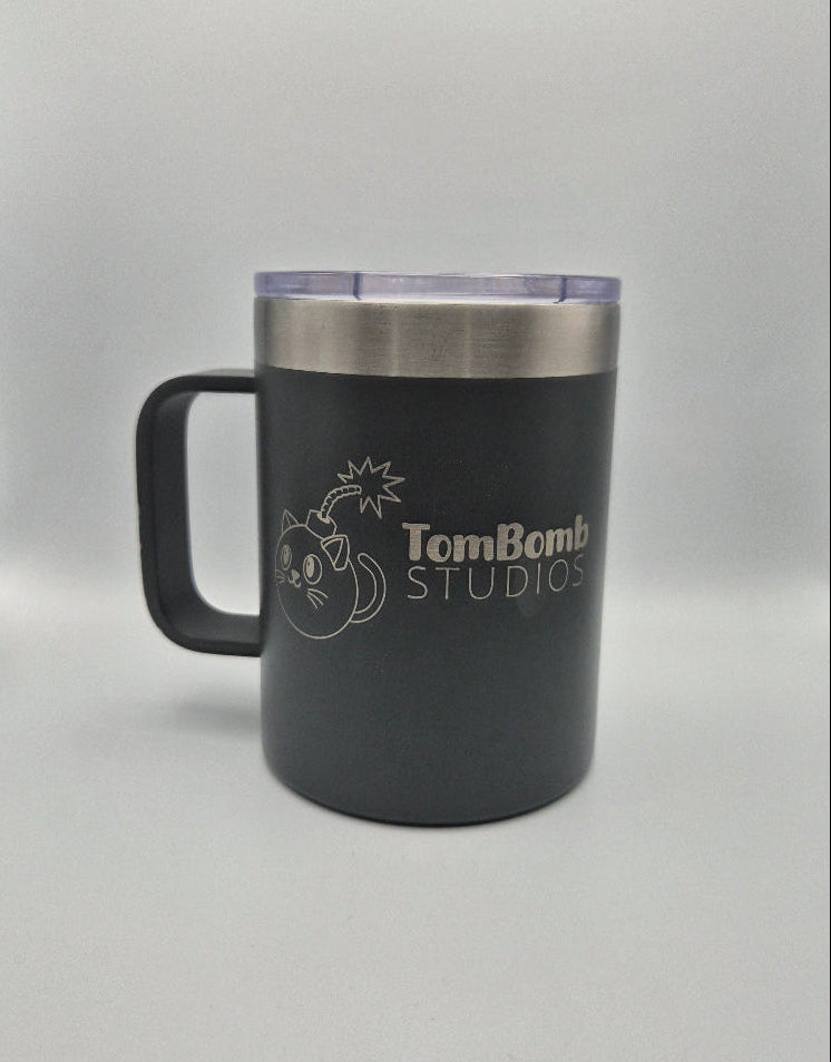 A matte black insulated travel mug with a stainless steel rim and a purple-tinted lid, featuring a white laser-engraved 'Tom Bomb Studios' logo on the front, set against a white studio backdrop.