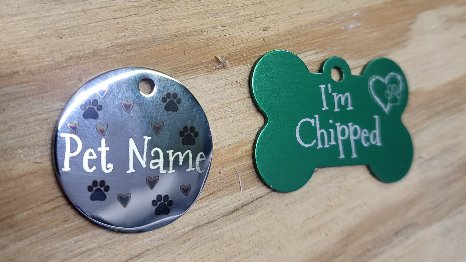 Two pet ID tags on a wooden surface: a round silver tag with paw prints and the text 'Pet Name,' and a bone-shaped green tag with 'I'm Chipped' and a small heart outline.